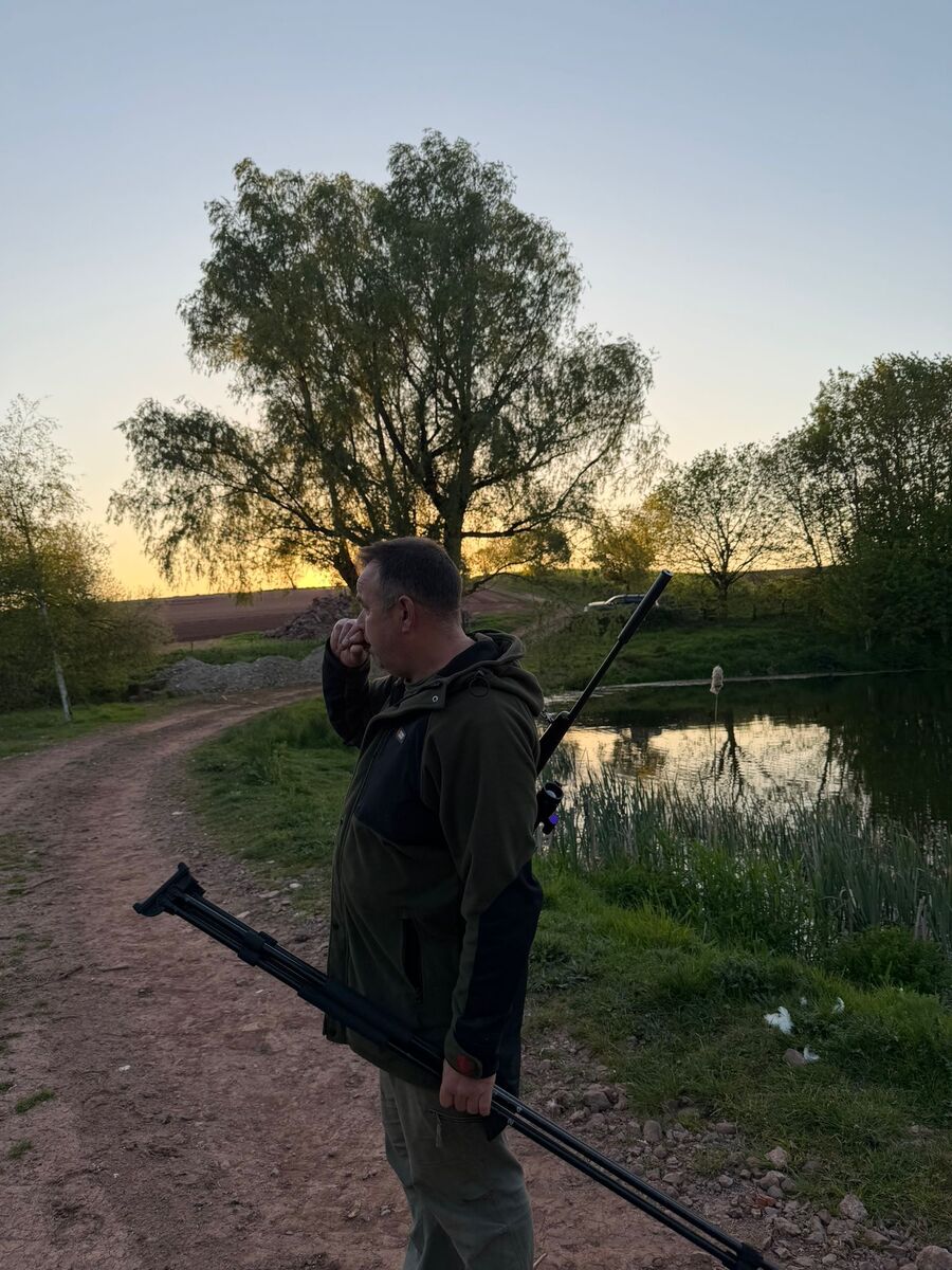 Julian Goodwin with rifle and shooting sticks beside a pond at dusk